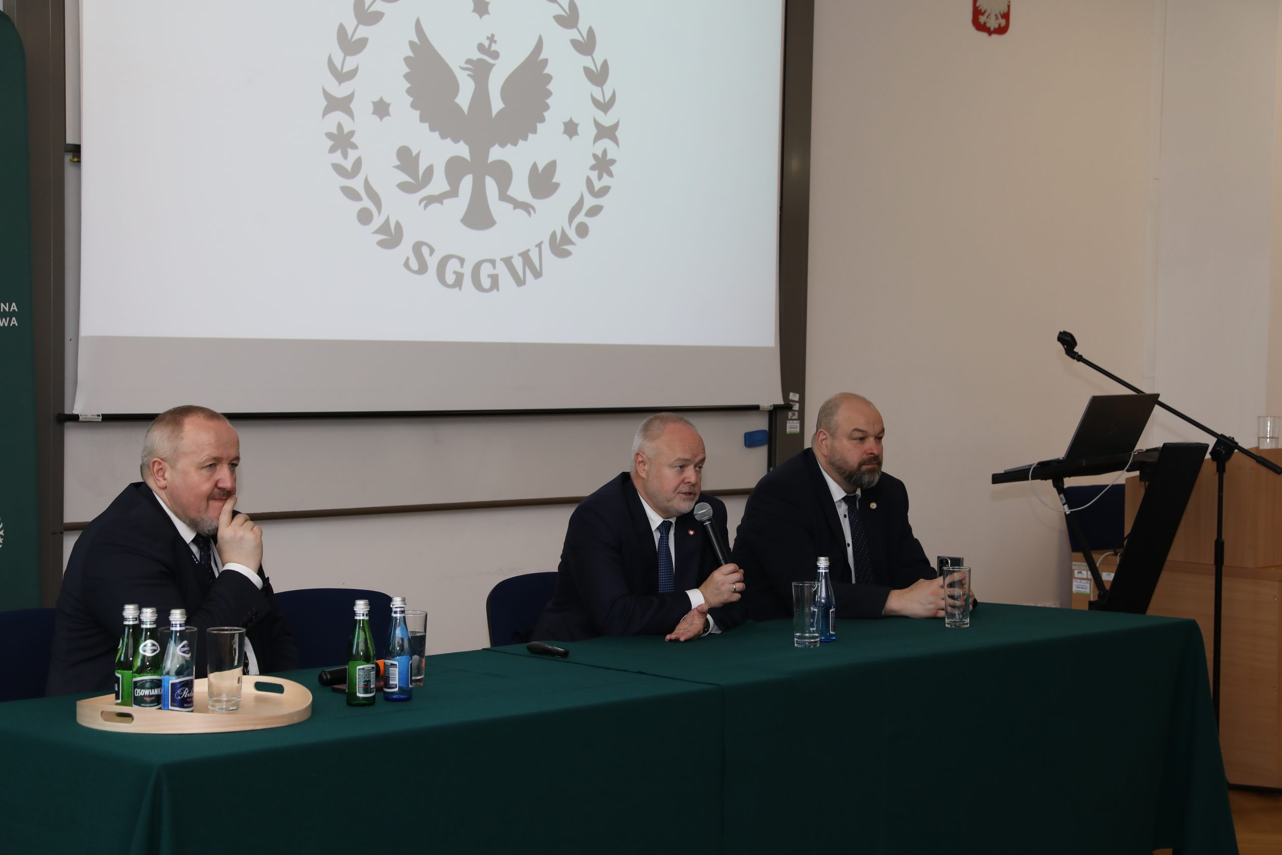 Trzej mężczyźni w garniturach siedzą przy zielonym stole konferencyjnym w SGGW
