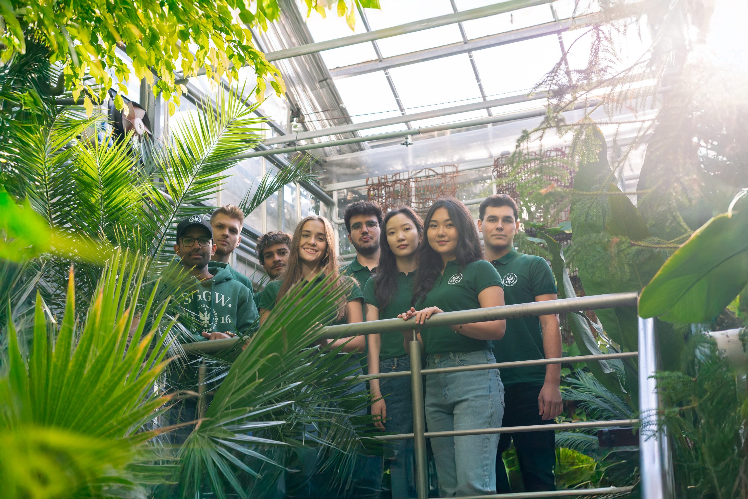 Grupa studentów SGGW w otoczeniu roślin w szklarni; stoją na metalowych schodach, uśmiechnięci, ubrani w zielone koszulki i bluzy z logo uczelni