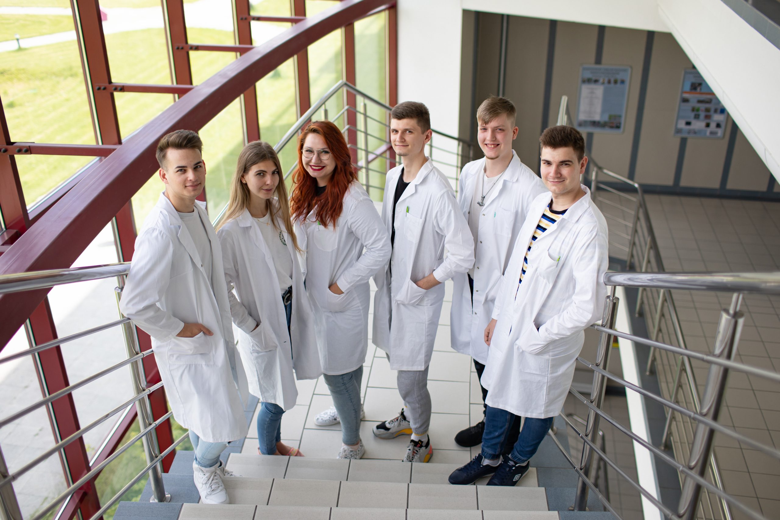 Grupa sześciu młodych osób w białych fartuchach laboratoryjnych stoi na schodach w budynku uczelni, patrząc w stronę obiektywu.