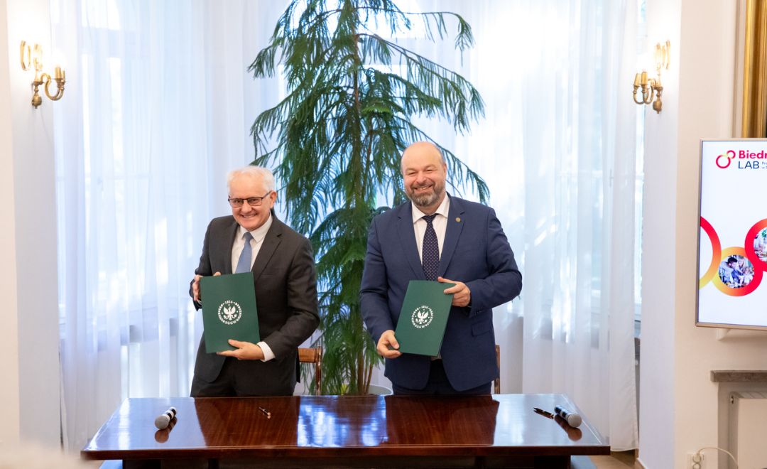 Dwaj mężczyźni w garniturach stoją przy stole, trzymając zielone teczki z godłem Szkoły Głównej Gospodarstwa Wiejskiego w Warszawie.