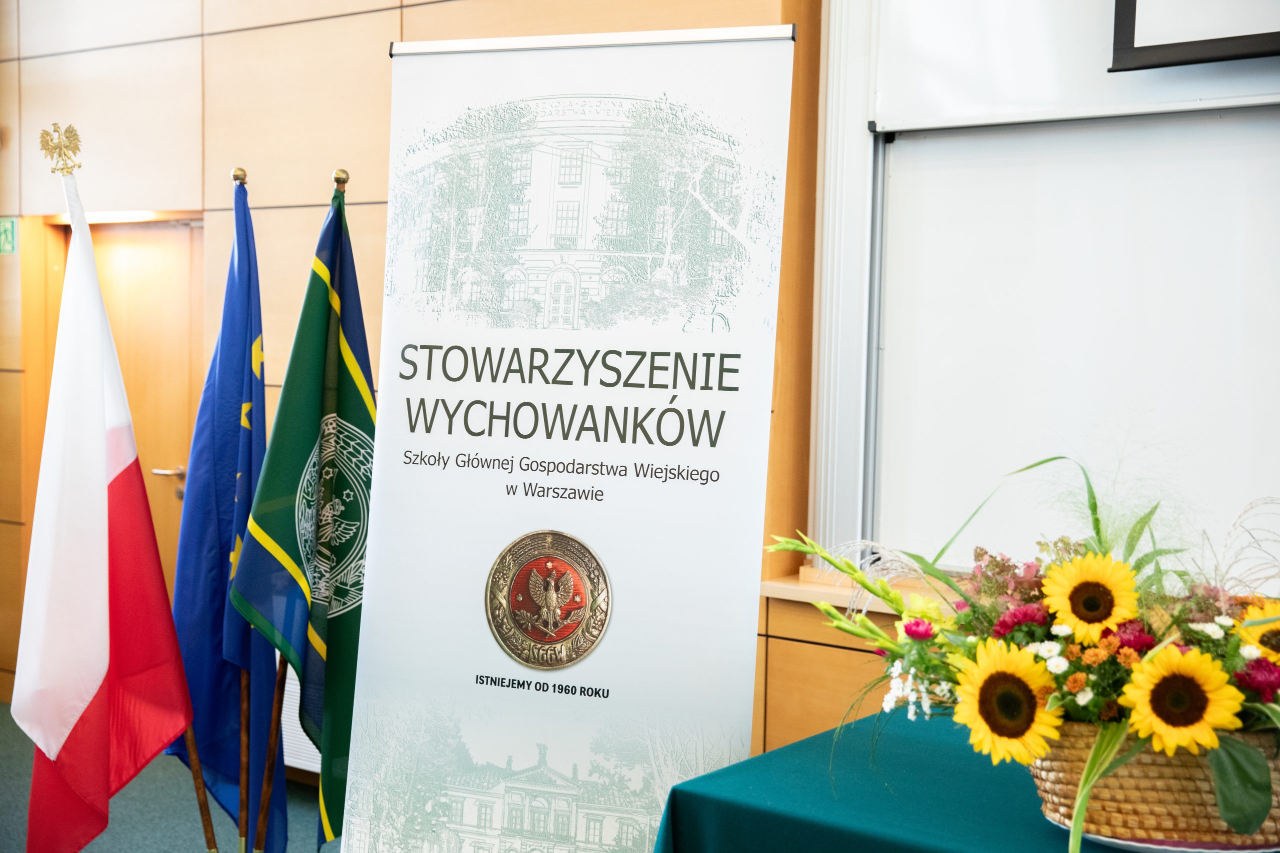 baner z napisem Stowarzyszenie Wychowanków SGGW, obok banera bukiet kwiatów i flagi