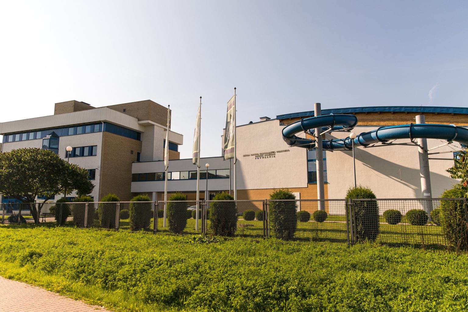 Kompleks obiektów sportowych SGGW, w tym charakterystyczny budynek pływalni z zewnętrzną, rurową zjeżdżalnią wodną w odcieniach niebieskiego. Nowoczesna bryła obiektu łączy jasne elewacje z elementami cegły i dużymi przeszkleniami.