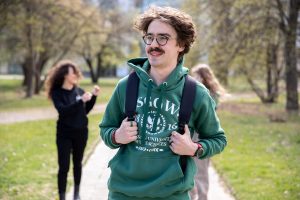Na zdjęciu widać młodego mężczyznę spacerującego po parku lub kampusie w słoneczny dzień. Ma na sobie zieloną bluzę z kapturem z logo SGGW oraz plecak. Uśmiecha się, patrząc lekko w bok. Charakterystyczne elementy to okulary i wąsy. W tle, lekko rozmyte, znajdują się dwie inne osoby oraz drzewa i alejka, co nadaje fotografii swobodny, studencki klimat.