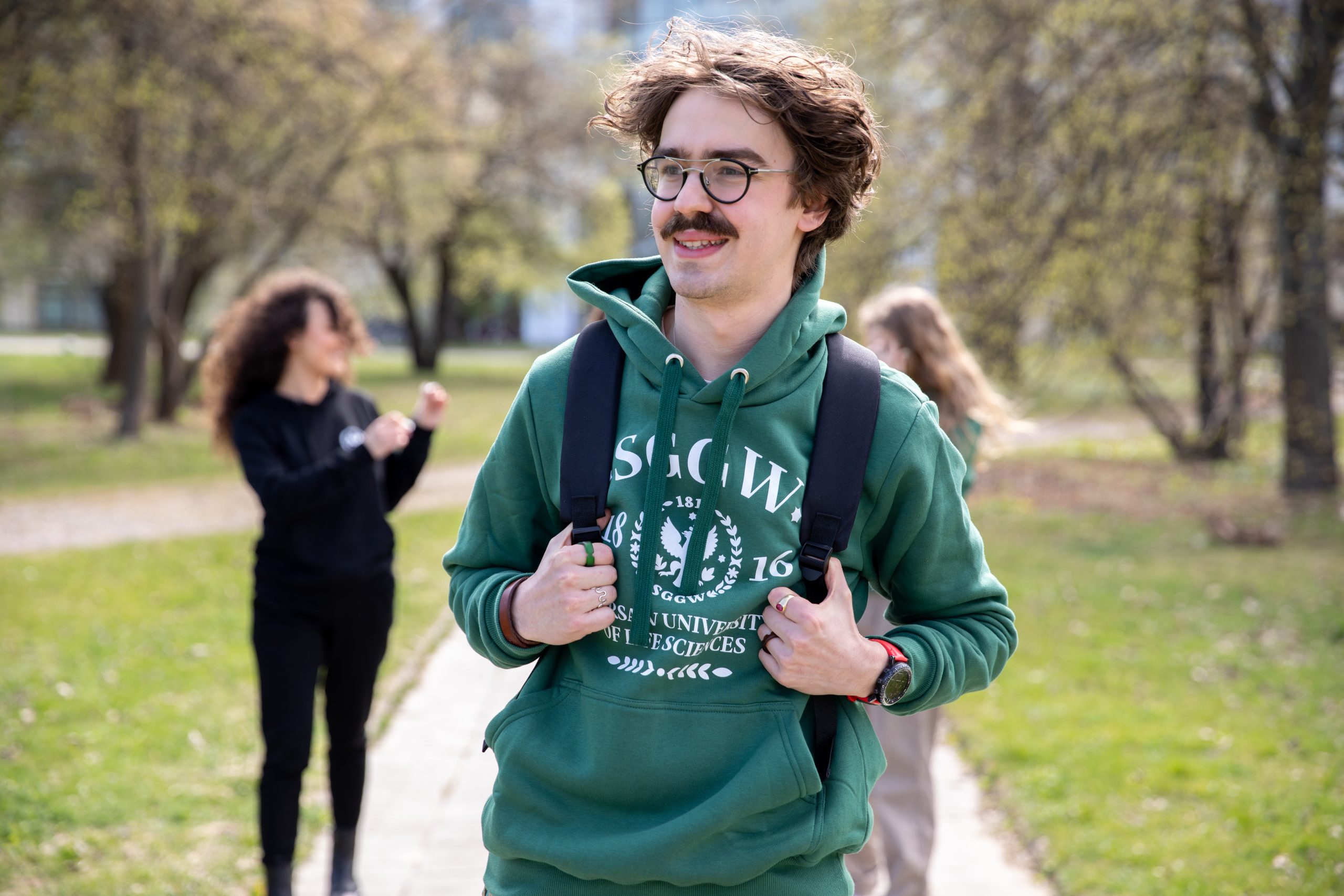 Na zdjęciu widać młodego mężczyznę spacerującego po parku lub kampusie w słoneczny dzień. Ma na sobie zieloną bluzę z kapturem z logo SGGW oraz plecak. Uśmiecha się, patrząc lekko w bok. Charakterystyczne elementy to okulary i wąsy. W tle, lekko rozmyte, znajdują się dwie inne osoby oraz drzewa i alejka, co nadaje fotografii swobodny, studencki klimat.