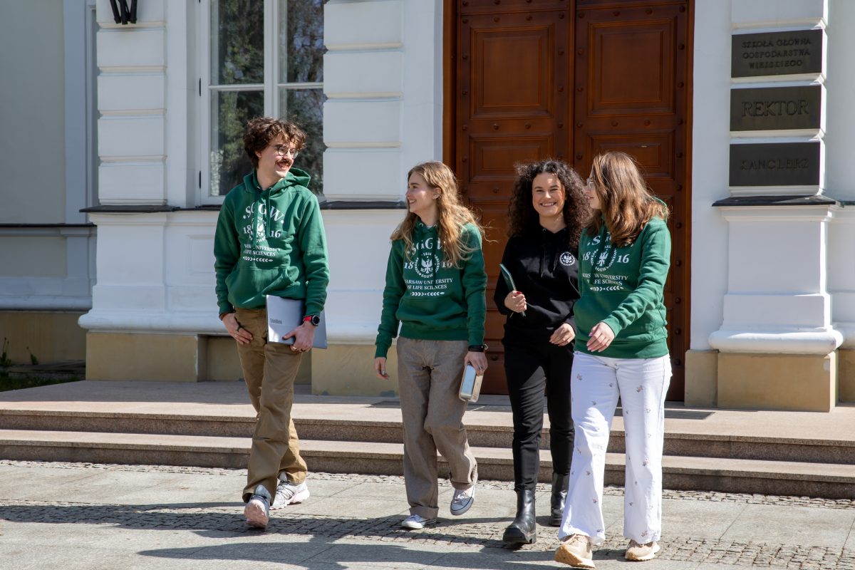 Na zdjęciu widać grupę czworga młodych osób wychodzących z reprezentacyjnego budynku uczelni. Trzy osoby mają na sobie zielone bluzy z logo SGGW, co wyraźnie wskazuje na związek ze społecznością akademicką. Jedna z osób ubrana jest na czarno i trzyma w ręku notes lub tablet.