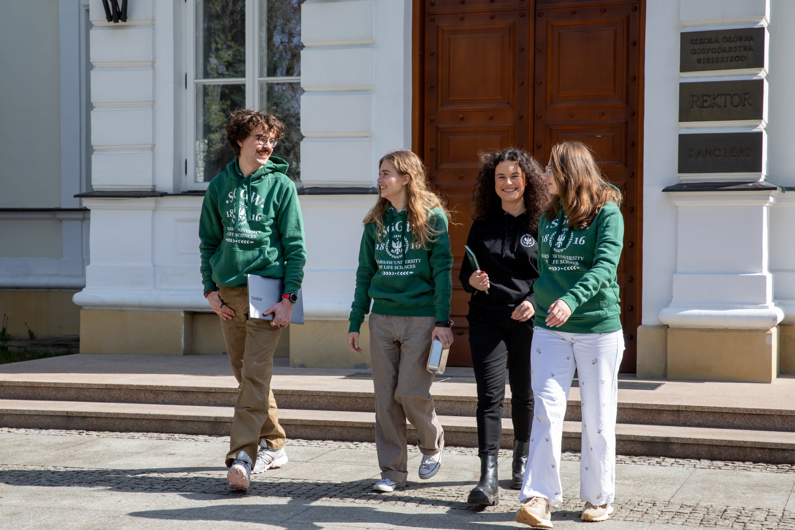 Na zdjęciu widać grupę czworga młodych osób wychodzących z reprezentacyjnego budynku uczelni. Trzy osoby mają na sobie zielone bluzy z logo SGGW, co wyraźnie wskazuje na związek ze społecznością akademicką. Jedna z osób ubrana jest na czarno i trzyma w ręku notes lub tablet.